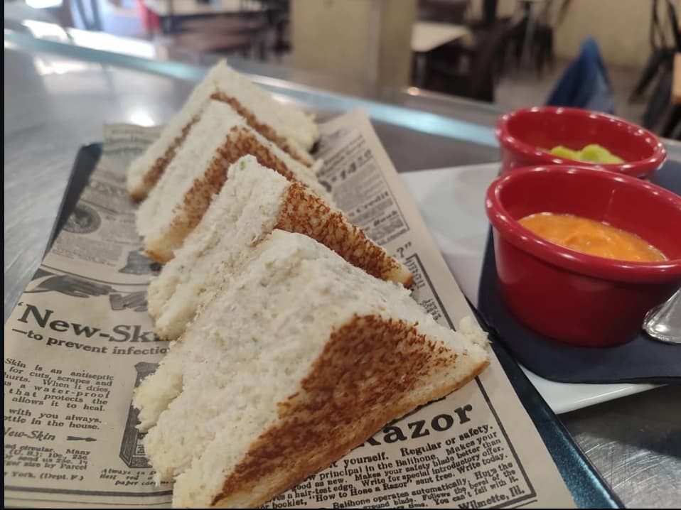Sándwiches triangulares tostados sobre papel de periódico acompañados de dos cuencos rojos con salsas.